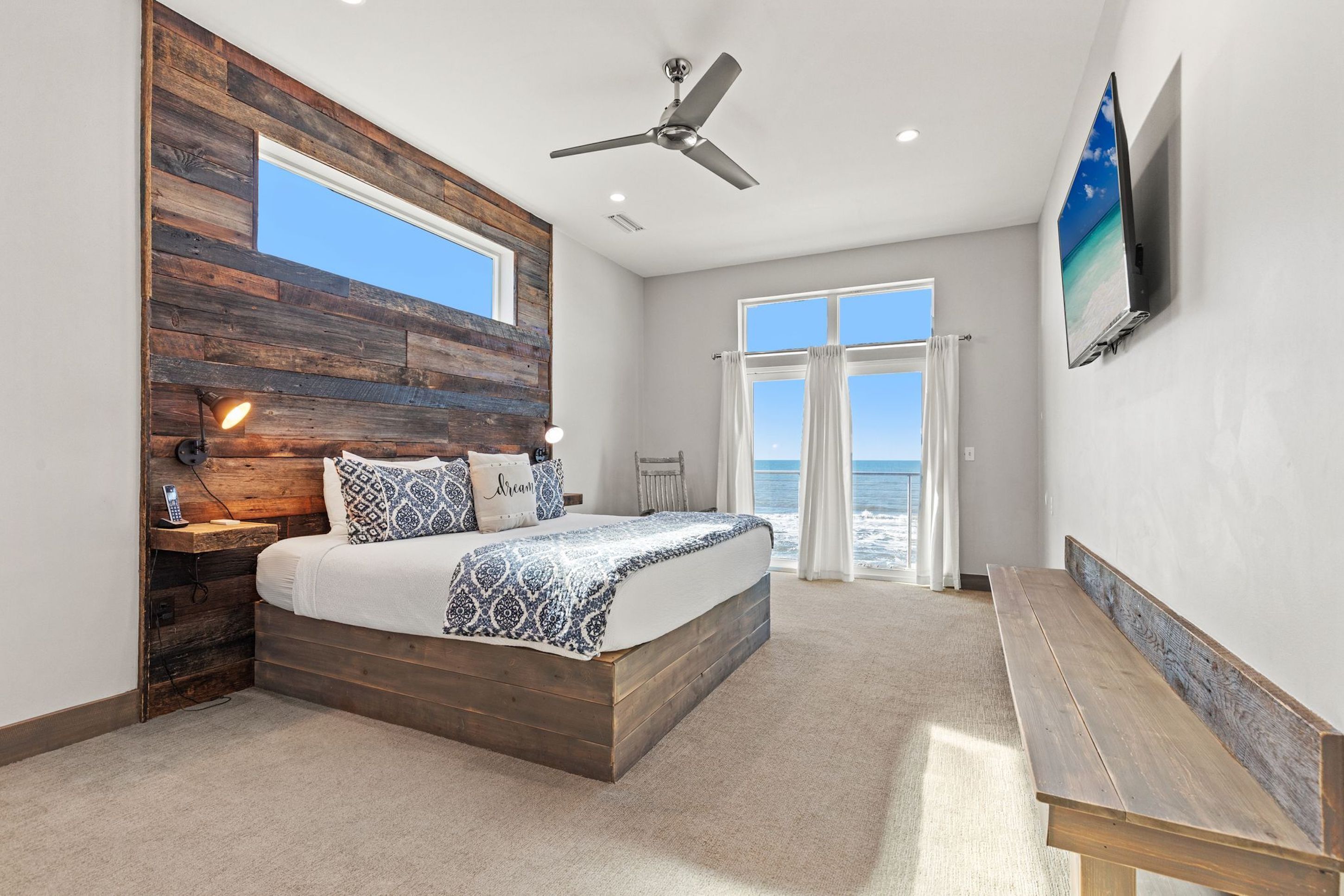 Quarto com cabeceira de madeira, cama com roupa de cama estampada, ventilador de teto, TV na parede e vista do oceano através de grandes janelas.