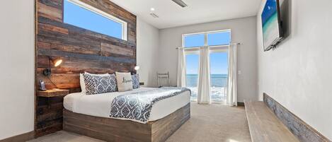 Quarto com cabeceira de madeira, cama com roupa de cama estampada, ventilador de teto, TV na parede e vista do oceano através de grandes janelas.