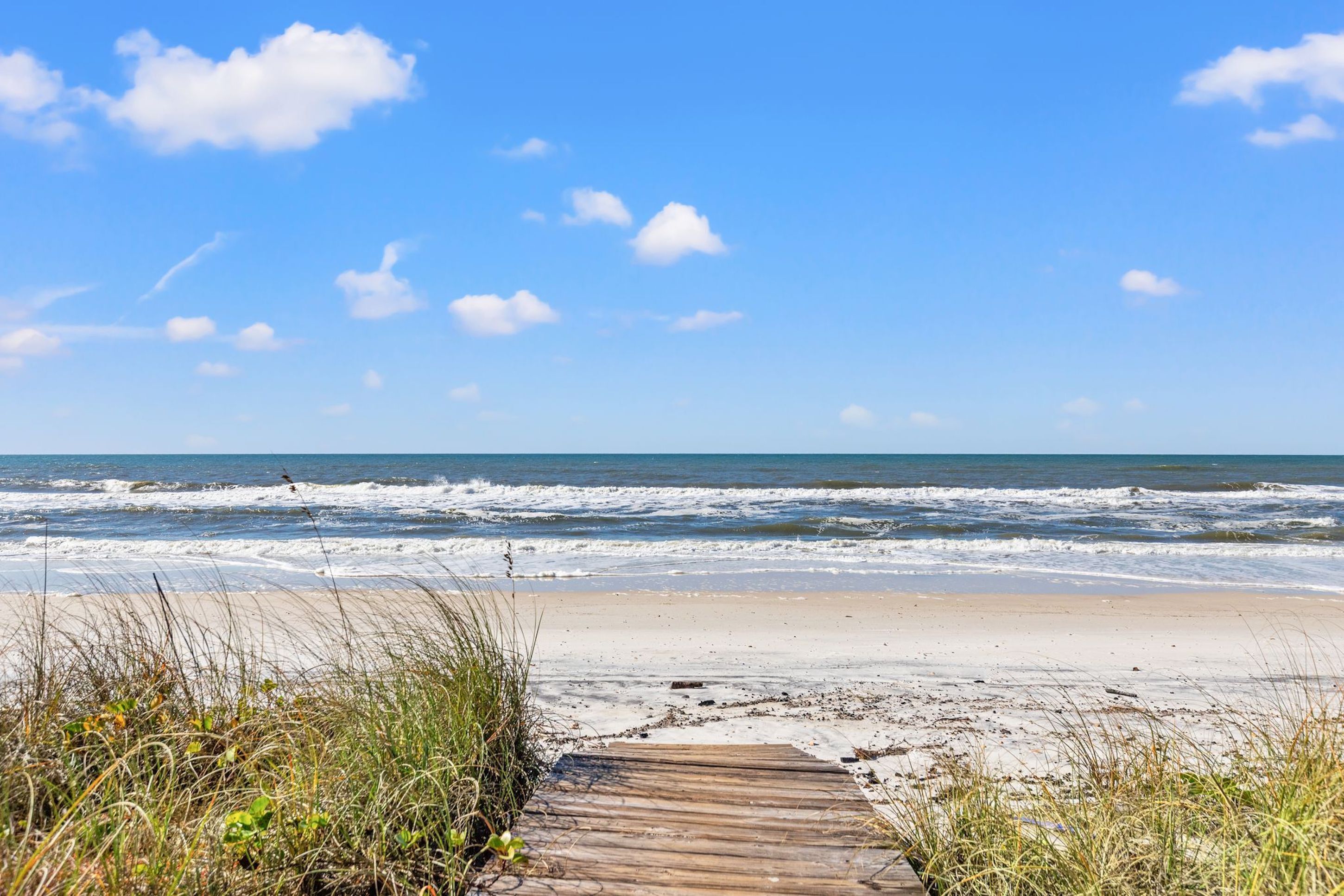 Ein hölzerner Gehweg führt zu einem Sandstrand, im Hintergrund schlagen Wellen und ein klarer, blauer Himmel erstreckt sich darüber.