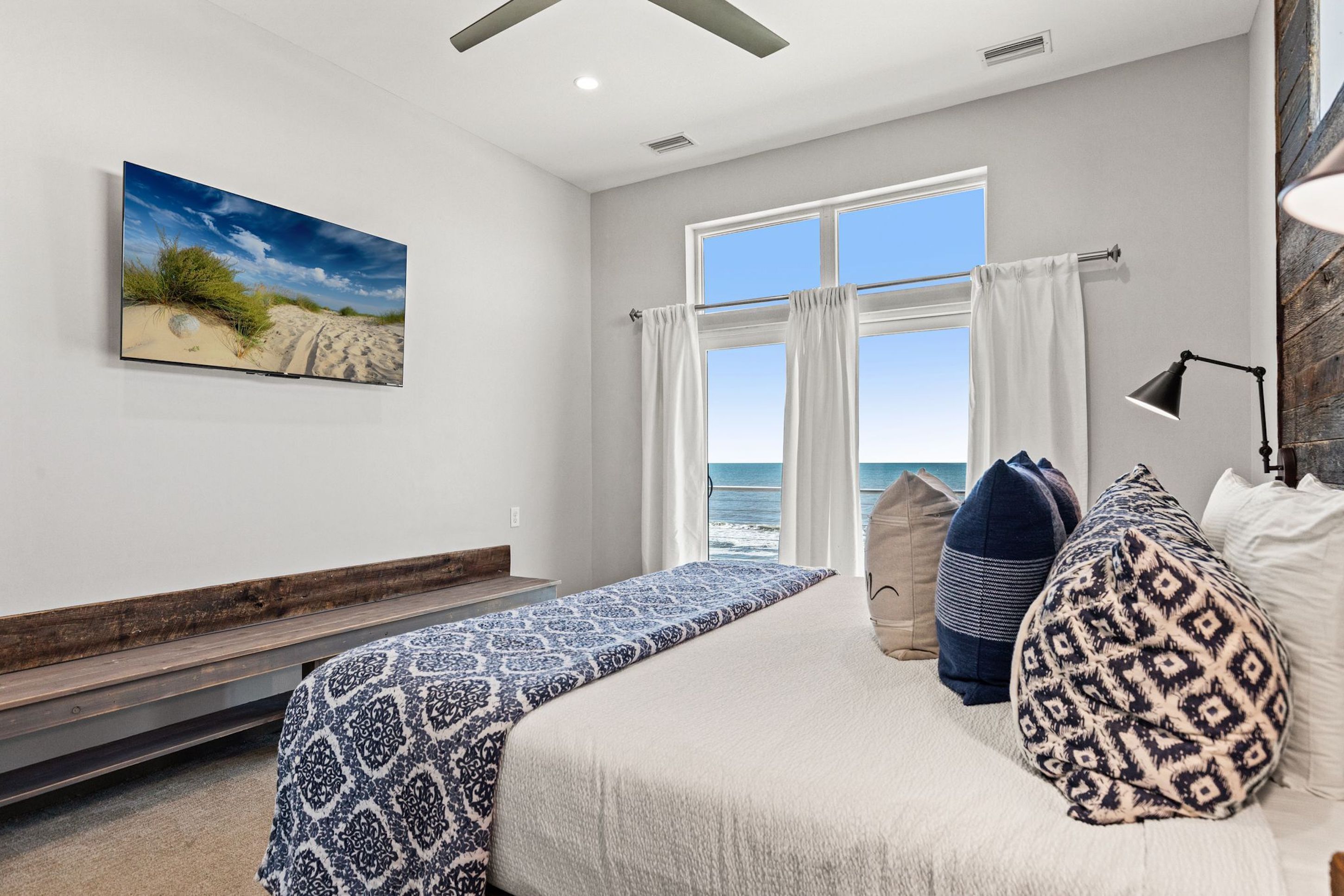 A bedroom with a large bed, a view of the ocean, a painting of a beach, and a wooden bench.