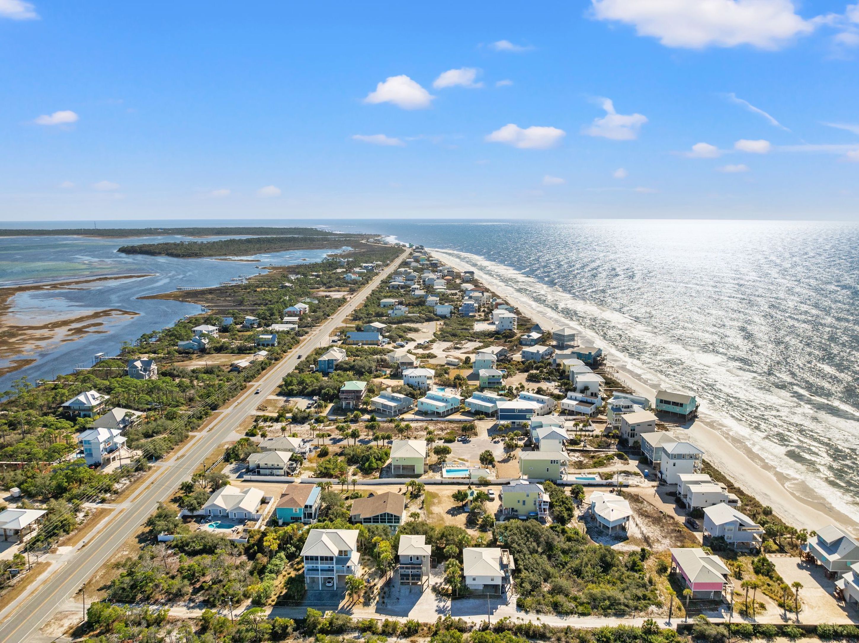 An aerial view of a coastal residential area with houses aligned along a road, a beach, and a clear sky.
