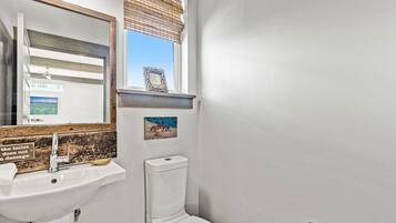 Un baño con lavamanos blanco, un espejo, inodoro y ventana con persianas.