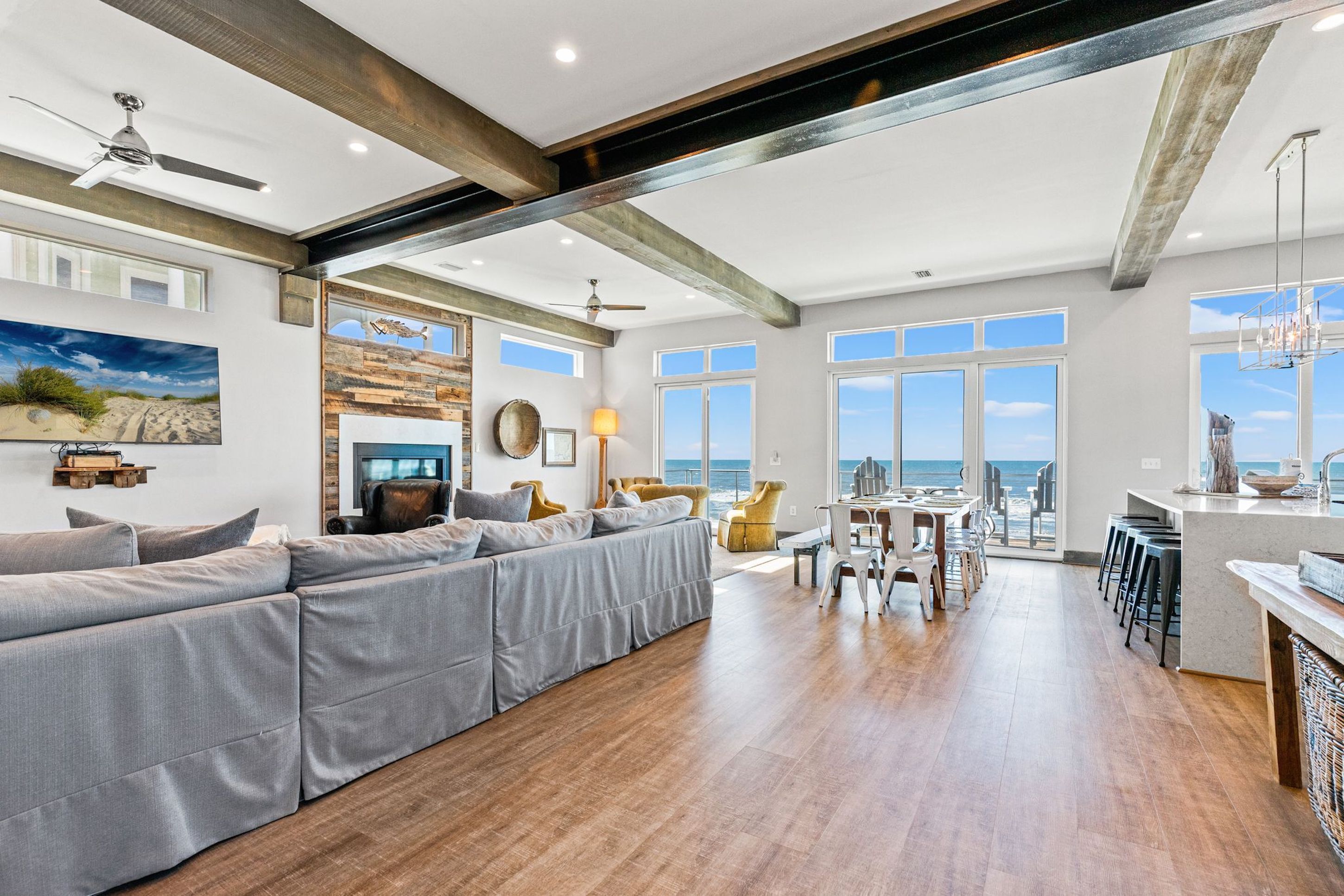 A spacious living area with a large gray sectional sofa, wooden flooring, and a view of the ocean through large windows.