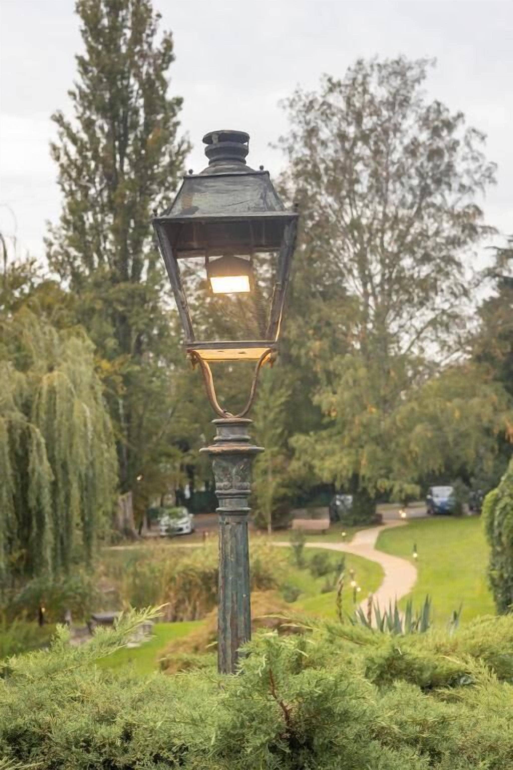 Un lampadaire traditionnel, installé dans un jardin, avec un sentier et des arbres en arrière-plan.