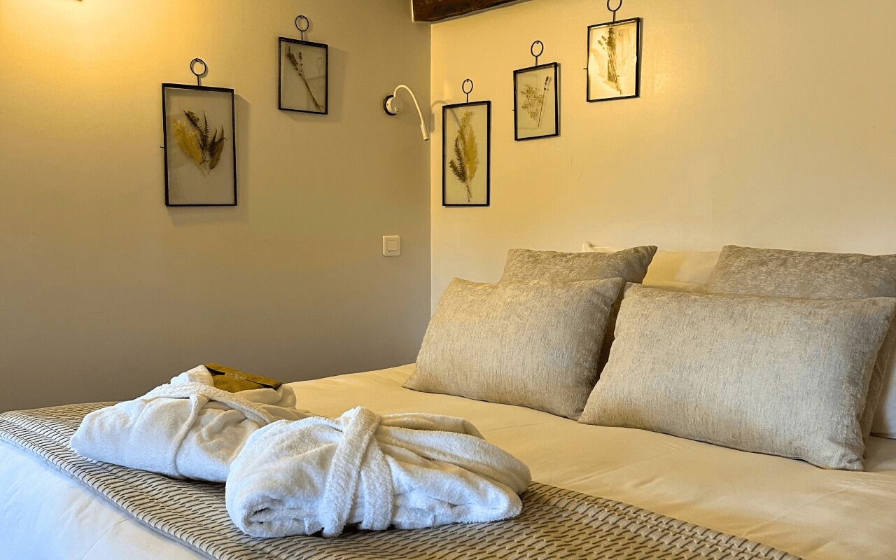 Una cama bien hecha con un albornoz blanco encima, una pared de color claro e impresiones botánicas enmarcadas.