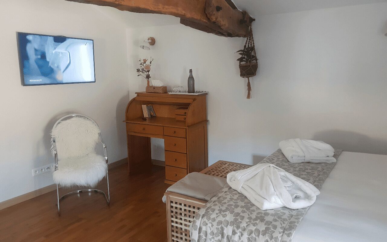 A bedroom with a wooden desk, a chair, a bed with towels, a hanging plant, and a wall-mounted TV.
