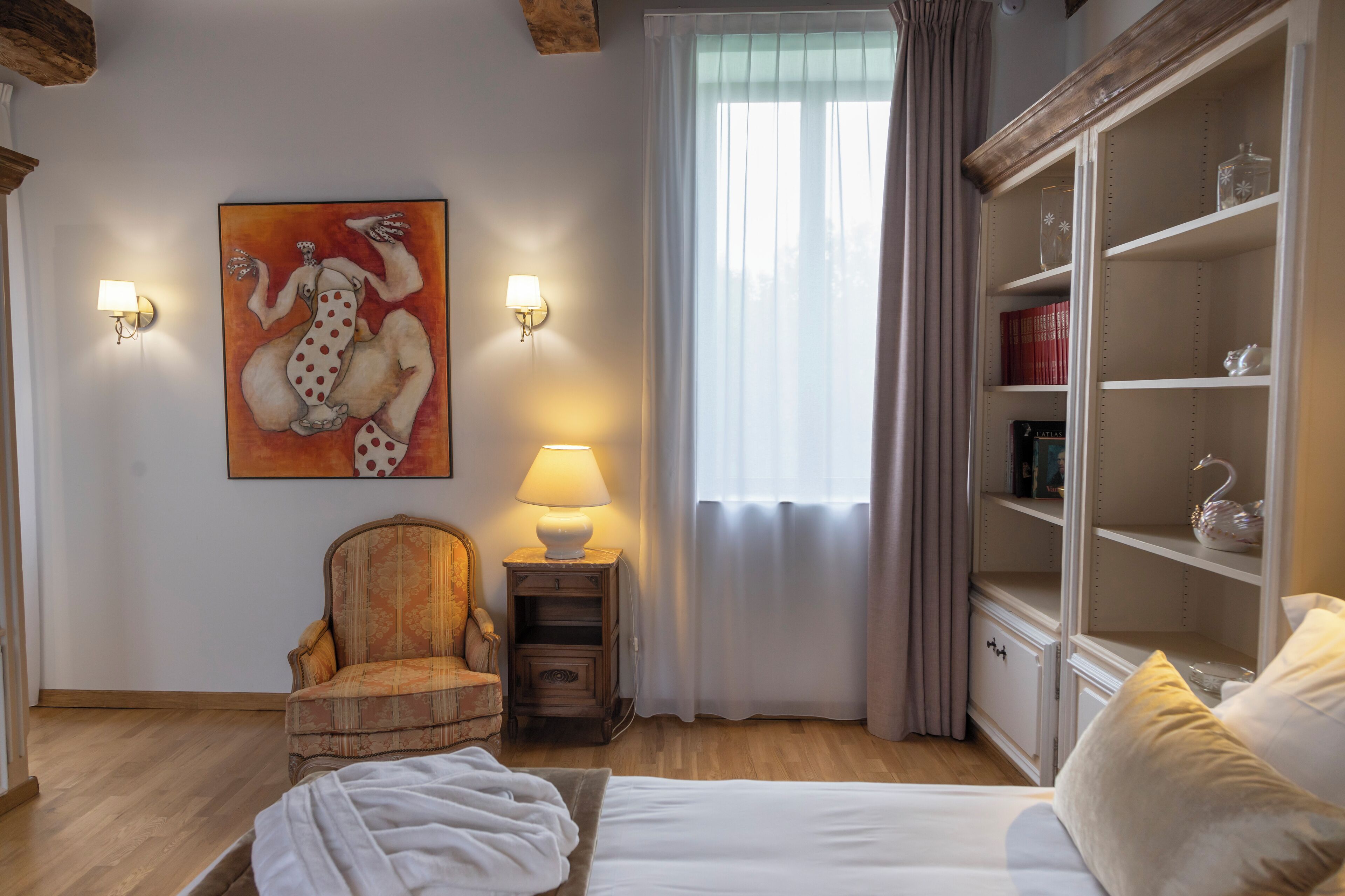 A bedroom with a bed, a chair, a nightstand with a lamp, a wardrobe, and a painting on the wall.