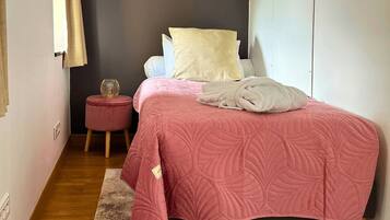 Un dormitorio con una cama, una mesita redonda pequeña, un taburete rojo y un armario blanco.