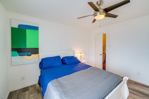 Ein Schlafzimmer mit einem Bett, einem Deckenventilator, einem Nachttisch mit Lampe und einer Tür zu einem weiteren Raum.