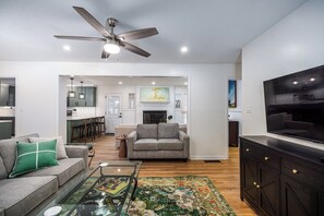 A modern living room with a grey sofa, a coffee table, and a TV on a black cabinet. There is a fireplace and a kitchen in the background. - 6.5 Acres - Remodeled Private Ranch. EV Charger. Fire pit. Comfy and calm. (Lilburn)