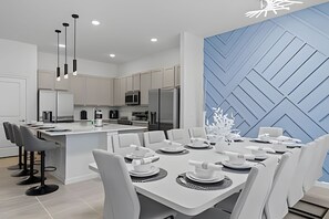 A modern kitchen with white cabinets, a central island, and a dining area with white chairs and a table set for six. - Luxurious 9BR Paradiso | Pool, Cinema & Game Room (Orlando)