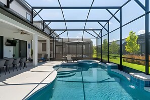 A modern outdoor pool area with a glass enclosure, a seating area with chairs and a table, and a view of the surrounding greenery. - Luxurious 9BR Paradiso | Pool, Cinema & Game Room (Orlando)
