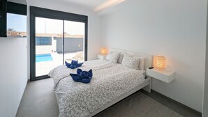 Un dormitorio con cama, mesita de noche, lámpara, luz en el techo y vista a una piscina a través de una puerta corredera.