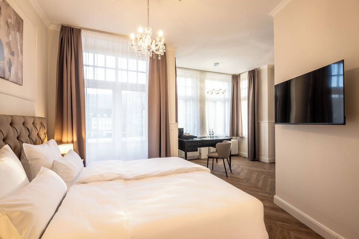 A hotel room with a large bed, a desk, a chair, and a TV on the wall.