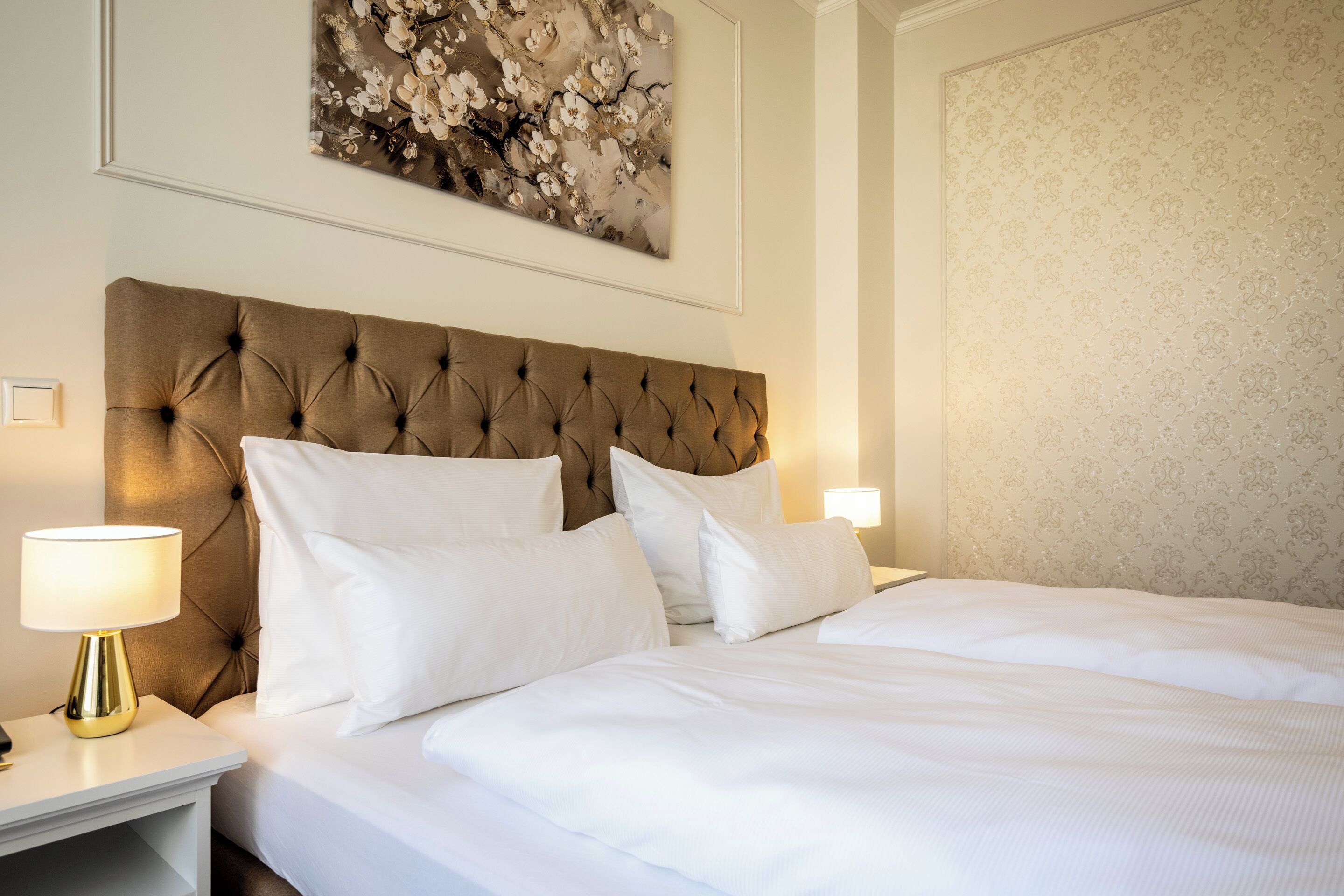 A neatly made bed with white linens and a tufted headboard, a bedside lamp, and a floral artwork above the bed.
