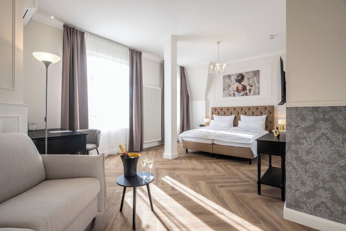 A hotel room with a bed, a sofa, a desk, a small table with a bottle and glasses, and a large window with curtains.