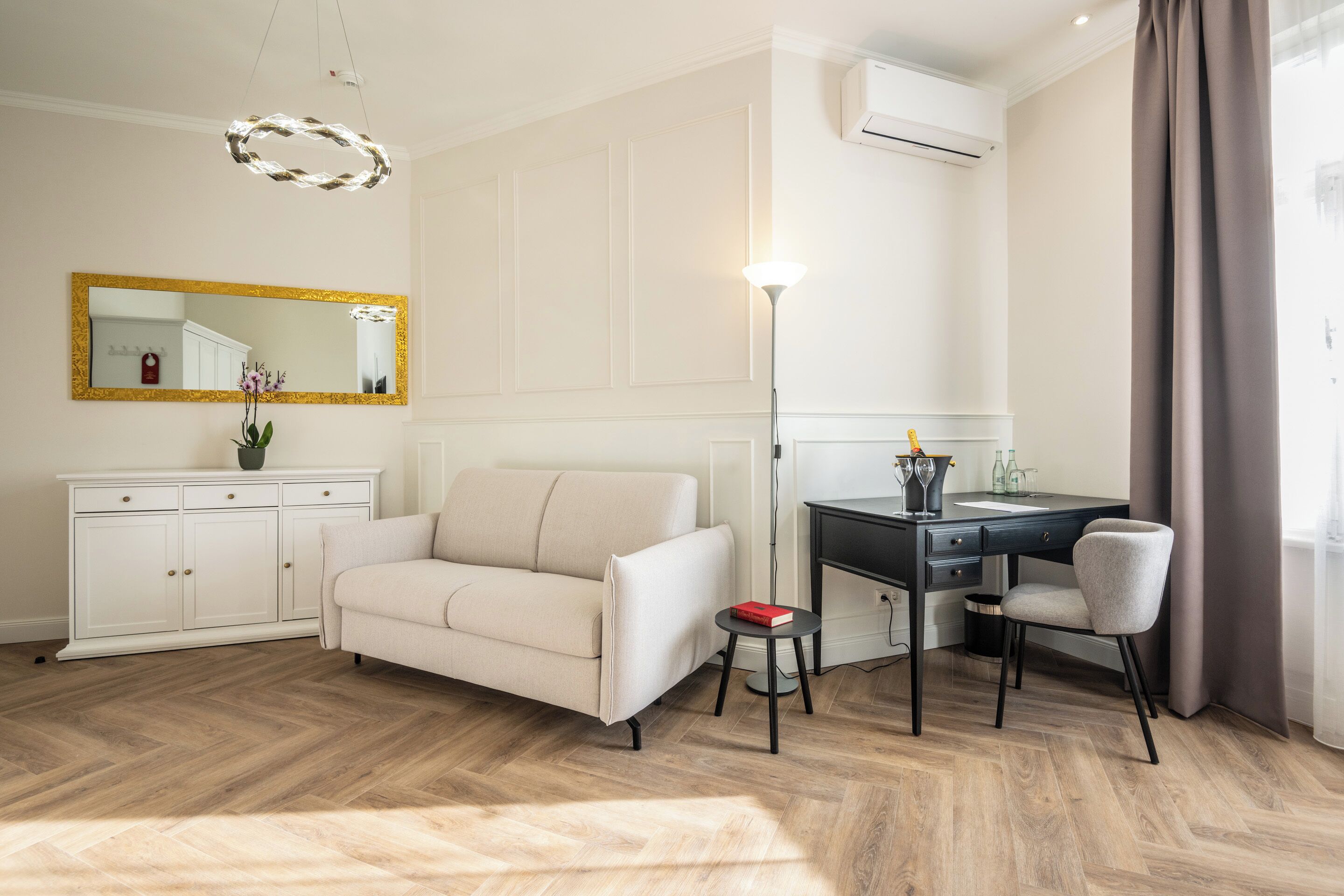 A modern room with a sofa, a desk, a chair, a small table, a mirror, and a hanging light fixture.