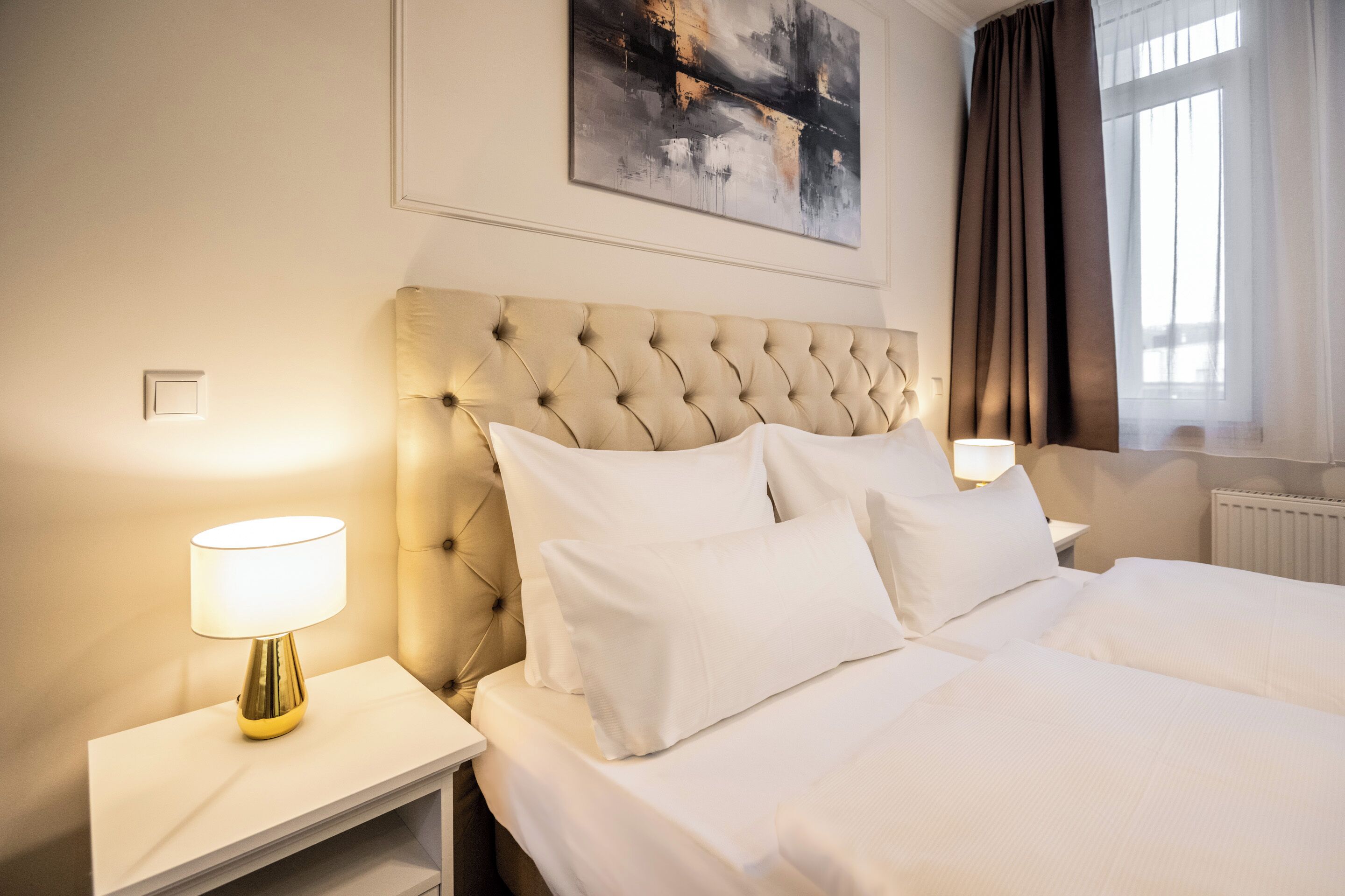 Una habitación de hotel con cabecero capitonado, ropa de cama blanca, una lámpara de noche y un cuadro en la pared.