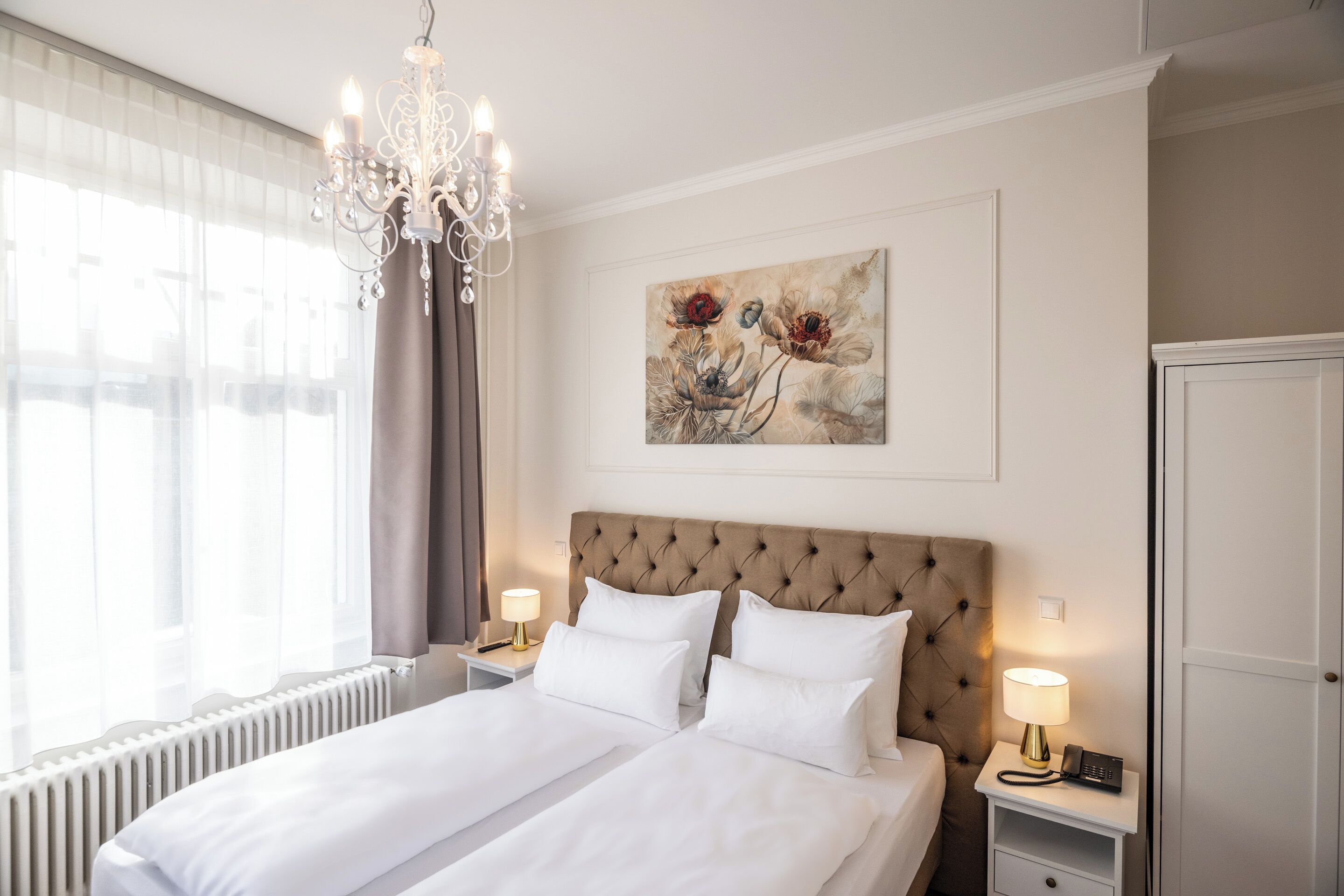 A hotel room with a large bed, two bedside tables, a chandelier, a radiator, and a painting on the wall.