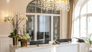 A hotel reception desk with a modern design, featuring a chandelier, a large window with curtains, and a sign that reads "HOTEL ALEX SEIT 2025".