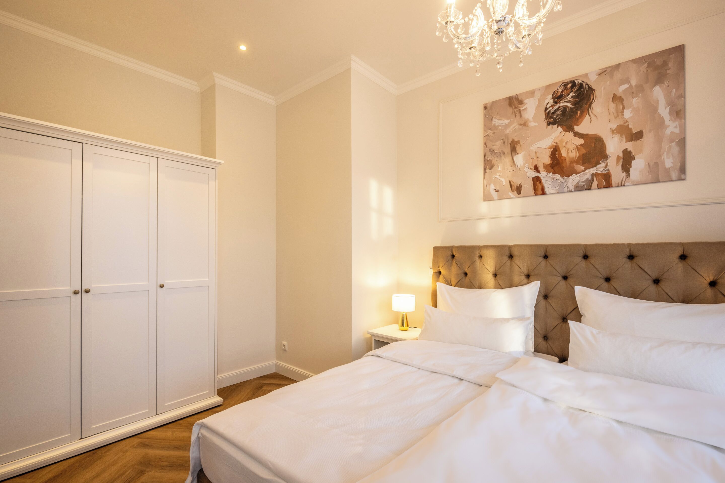 A bedroom with a large bed, a nightstand with a lamp, a wardrobe, and a painting on the wall.