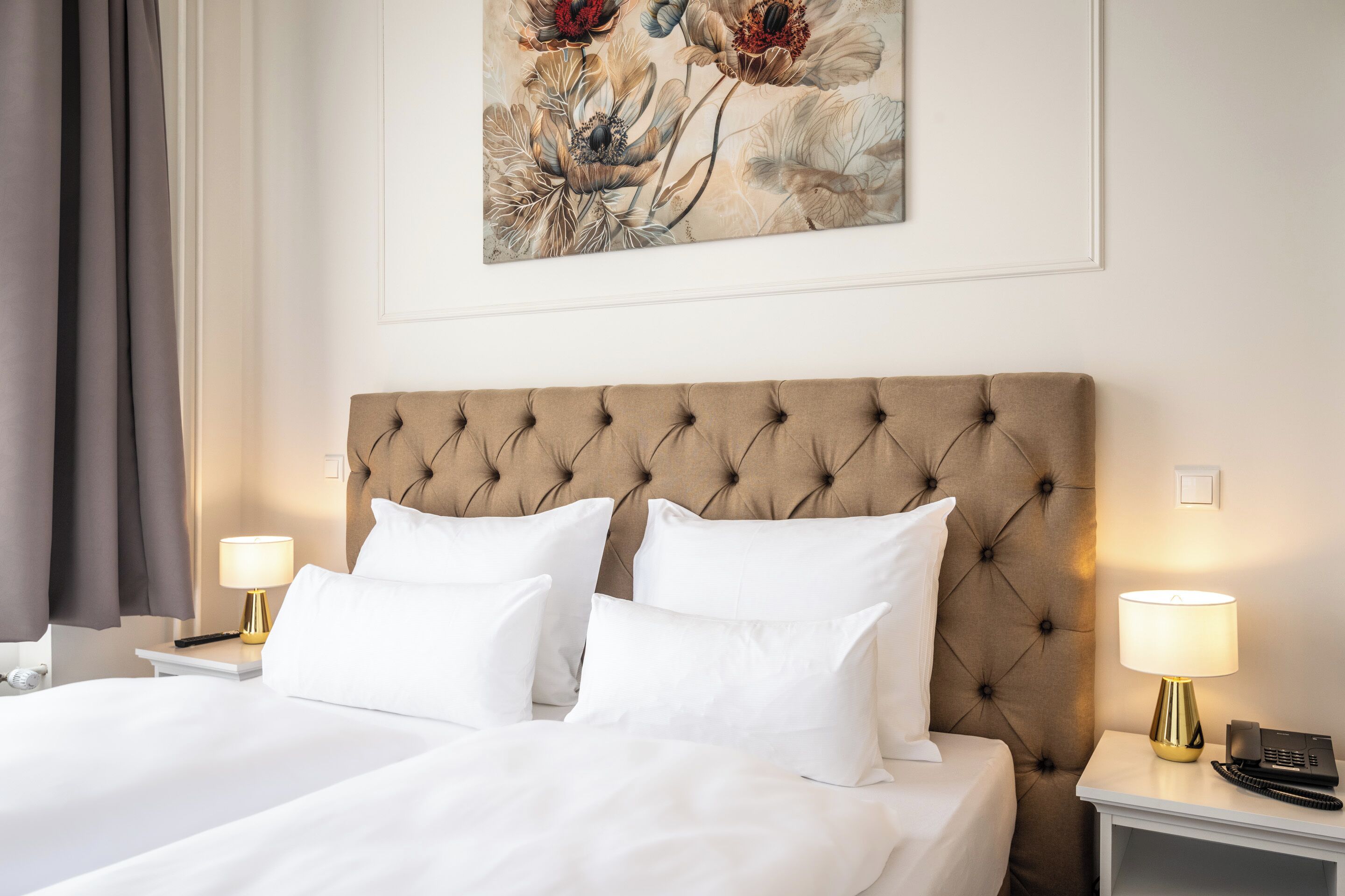 Habitación de hotel con cabecera capitonada, dos camas blancas, un cuadro en la pared y lámparas de noche.