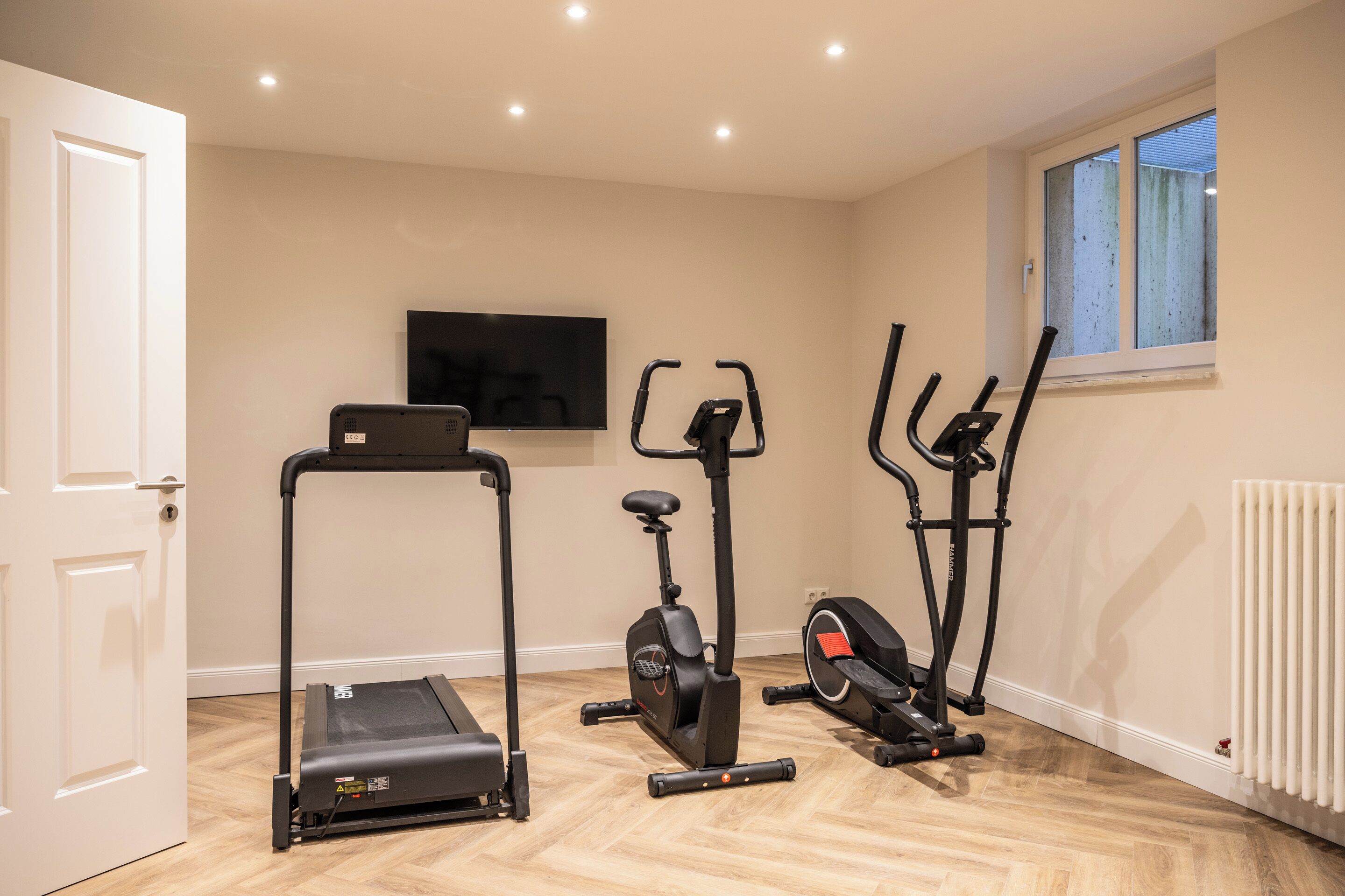 A room with a treadmill, elliptical, and stationary bike, a TV on the wall, and a window with blinds.