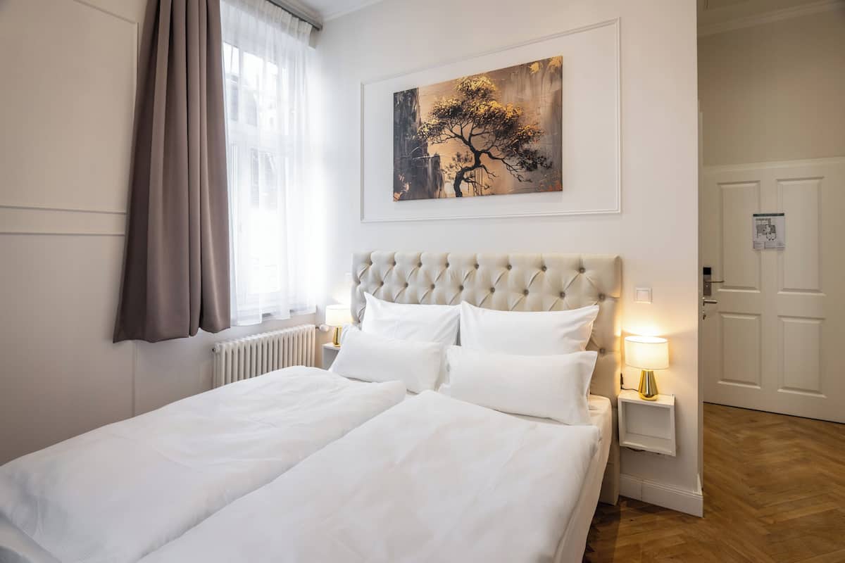 A hotel room with a large bed, a headboard, a nightstand with a lamp, a window with curtains, and a painting on the wall.
