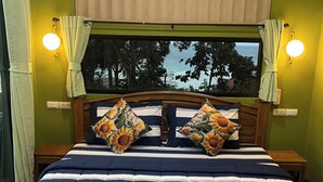A bed with a striped blue and white comforter, two pillows with sunflower designs, a wooden headboard, and a view of the ocean through a window with curtains. - The View Hostel (Ko Phi Phi)
