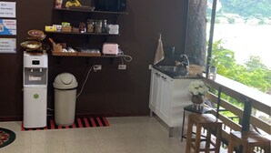 A room with a kitchenette, a dining area with wooden chairs, a large window with a view of greenery, and a wall-mounted coffee-themed artwork. - The View Hostel (Ko Phi Phi)