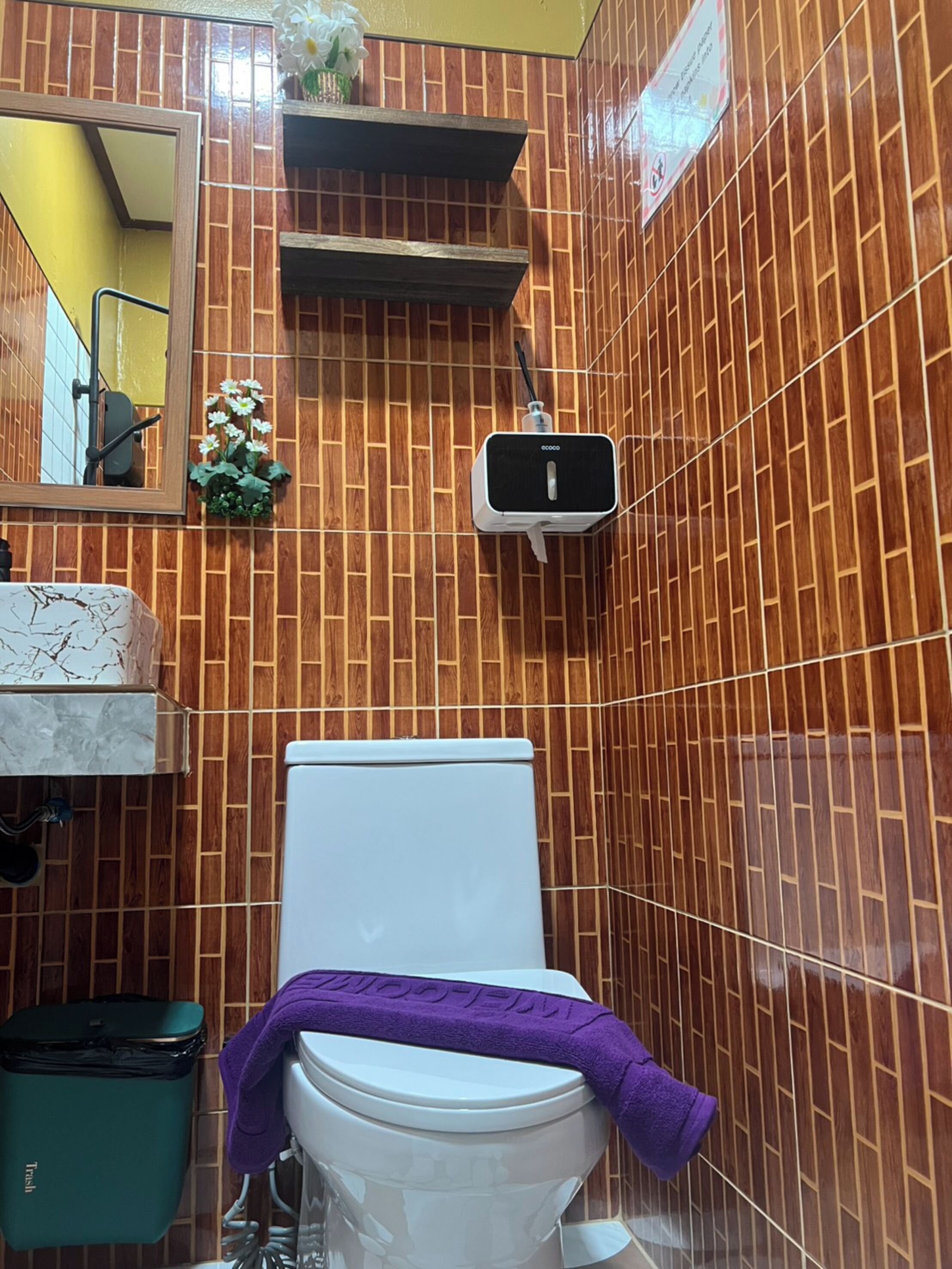 Ein Badezimmer mit Toilette, einem Mülleimer, einem Regal mit Pflanze und einem an der Wand befestigten Lufterfrischer.
