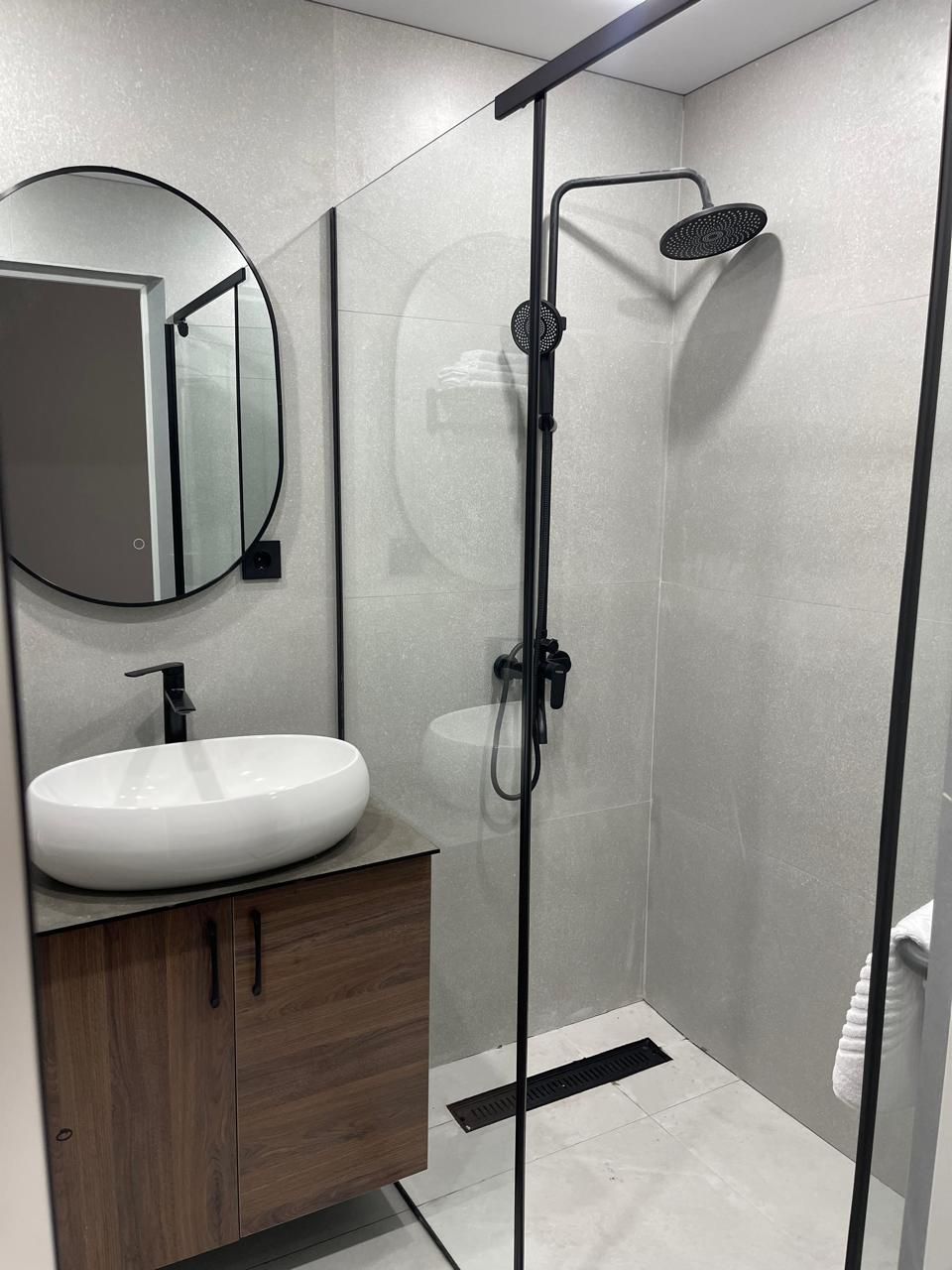 A modern bathroom with a round mirror, a white sink, wooden cabinets, and a glass-enclosed shower area.