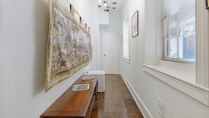 A hallway with a wooden bench, a large tapestry, a chandelier, and a window with blinds. - The Master Suite - A spacious suite inspired by Charleston’s grace and charm (Charleston)