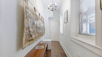 A hallway with a wooden bench, a large tapestry, a chandelier, and a window with blinds.