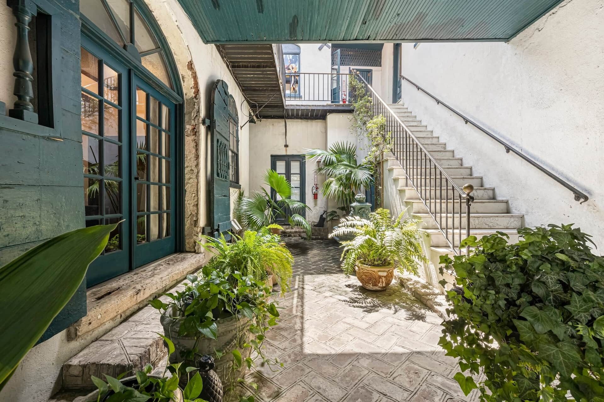A narrow courtyard with a staircase leading to an upper level, surrounded by potted plants and a rustic stone floor.