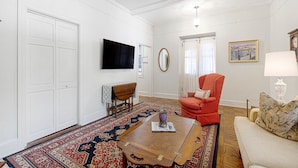 A living room with a large wooden coffee table, a red armchair, a beige sofa, a television, and a patterned rug. - The Master Suite - A spacious suite inspired by Charleston’s grace and charm (Charleston)
