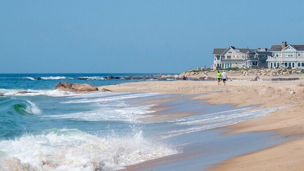 Uma praia com ondas, uma casa grande e céu claro.