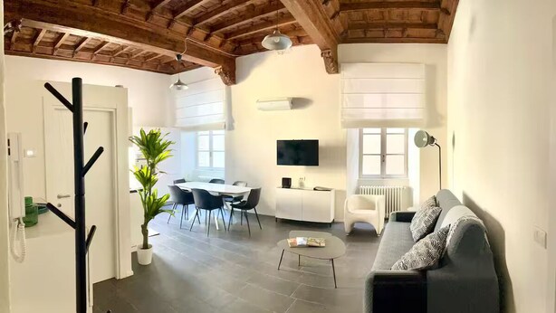 A modern living room with a sofa, coffee table, and dining area with chairs. There is a TV mounted on the wall, a plant, and a ceiling with exposed beams. - "La Dama alla Finestra," charming apartment in the historic center of Como. (Como)