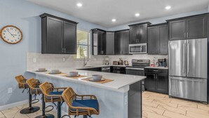 A modern kitchen with dark cabinets, a stainless steel refrigerator, and a central island with bar stools. - Ohana Memories At Championsgate (Davenport)