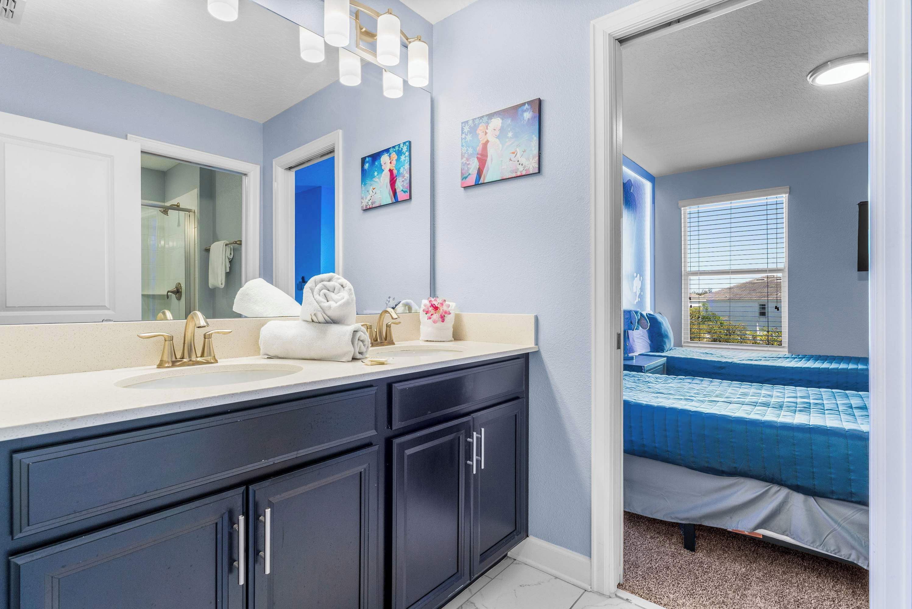 A bathroom with a large mirror, a sink, and a toilet. There is a bed with a blue bedspread in the adjacent room.
