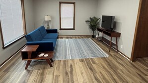 Una sala de estar con un sofá azul, una mesa de centro de madera, un televisor sobre un mueble bajo, un tapete estampado y una planta en maceta.