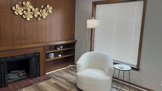 Una sala de estar con chimenea, paneles de madera, un sillón blanco, una mesita auxiliar y un ventanal con persianas.