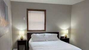Un dormitorio con una cama, dos mesitas de noche con lámparas, una ventana con persianas, una luz de techo y una obra de arte colgada en la pared.