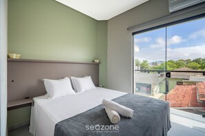 A modern hotel room with a large bed, a view of the city, and a balcony. - Apto moderno c/varanda em Campeche CRN101 (Florianópolis)
