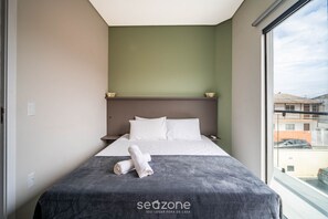 A hotel room with a bed, a window, and a view of buildings outside. - Apto moderno c/varanda em Campeche CRN101 (Florianópolis)