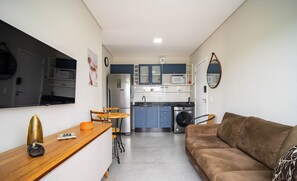 A compact living space with a kitchenette, a sofa, a dining area, and a TV mounted on the wall. - Apto moderno c/varanda em Campeche CRN101 (Florianópolis)