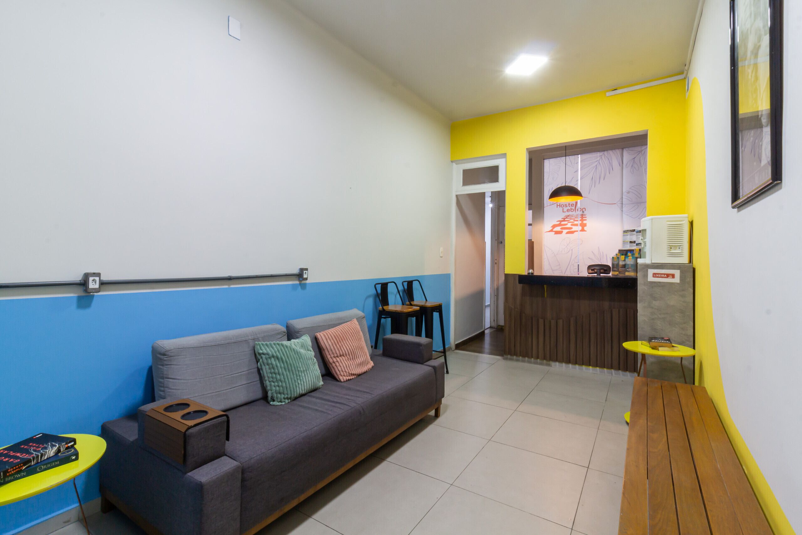 Un espace de vie comprenant un canapé, une petite table et un banc. Au fond, on aperçoit une kitchenette avec un comptoir et un luminaire suspendu.