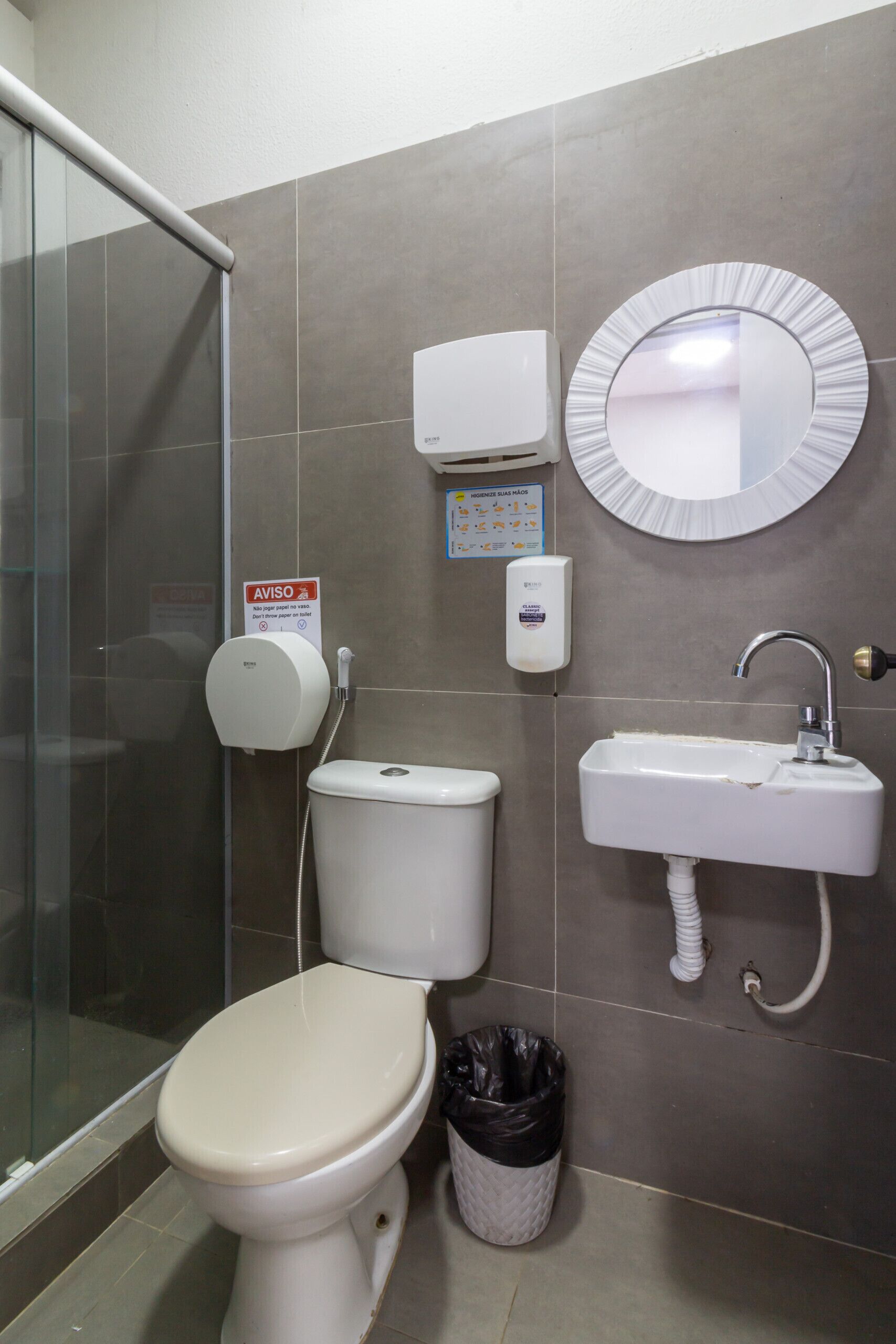 A bathroom with a toilet, a sink, a hand dryer, a trash bin, and a glass shower enclosure.