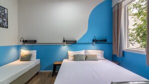 Une chambre d’hôtel avec un grand lit, un plus petit lit et un banc. Il y a une fenêtre avec des rideaux, une étagère fixée au mur avec des lampes et un tableau accroché au mur.