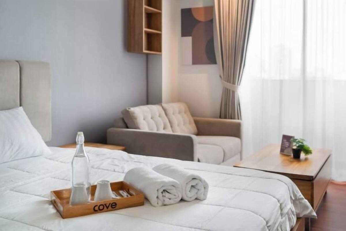 A hotel room with a bed, a side table, a sofa, and a window with curtains.